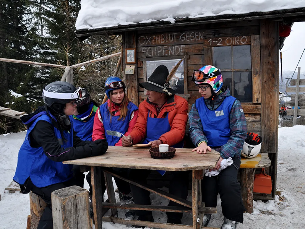 Gruppe sitzt vor einer Hütte mit einem handgeschriebenen Schild.